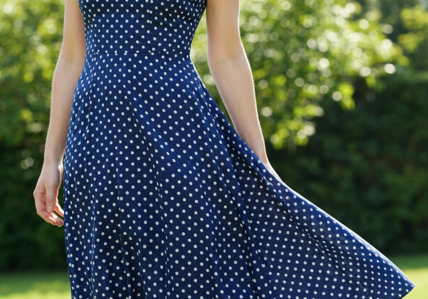 Kobieta w granatowej sukience w białe grochy stoi na tle rozmytej zieleni na zewnątrz w naturalnym świetle